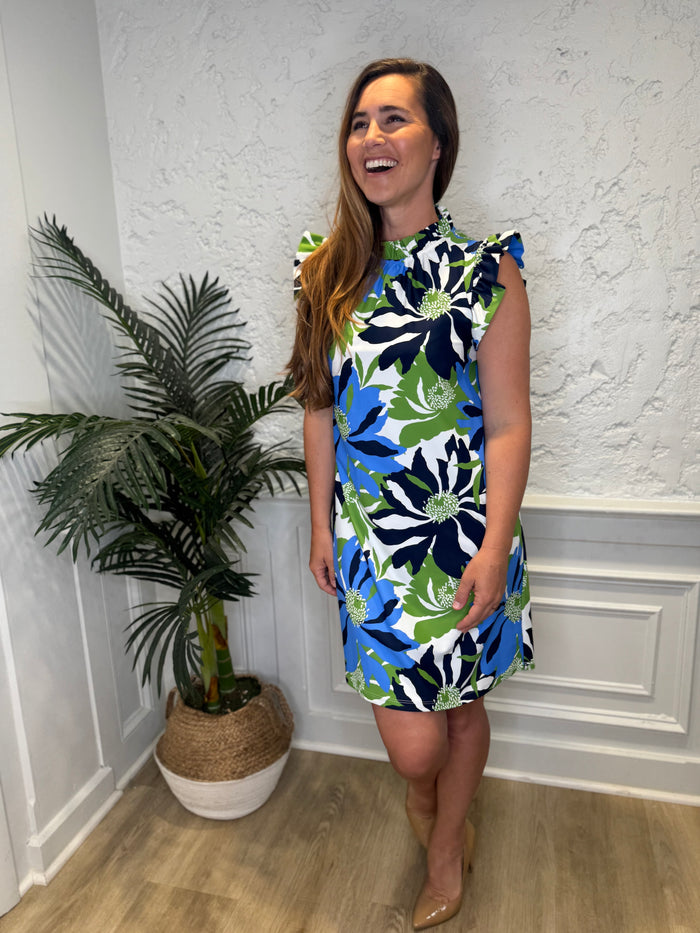 Smiling model in front of a white wall with a plant in the corner in the Jude Connally Shari Dress - Wild Blossom Blue. A short dress with flutter sleeves and collar, with a blue, green, and navy floral pattern