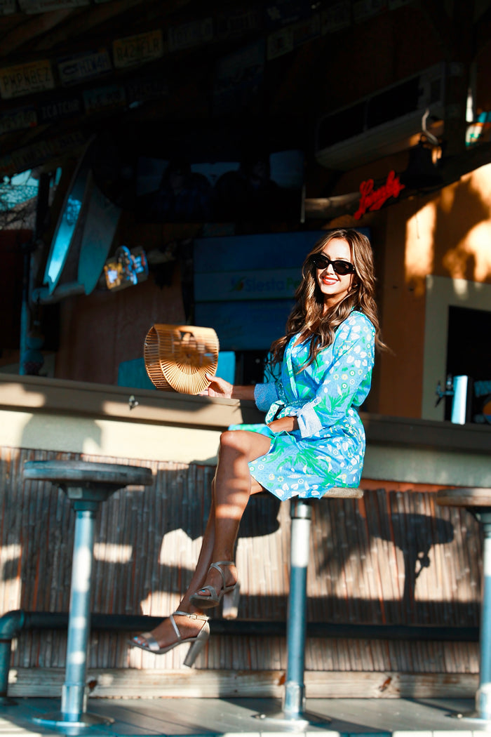 Model sitting at a beach bar in a knee length, green, blue, and white dress that has a belted waist, and contrasting cuff sleeves. She is wearing scrappy sandals and has a bamboo bag