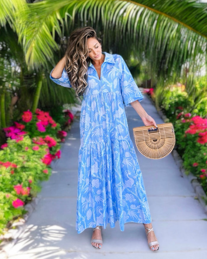 Model in a tiered maxi dress with a light blue and white floral and bird pattern with touches of green. V-neck with traditional collar and cuffed sleeves
