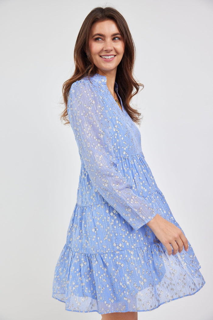 Model in a short, tiered dress with long sleeves and a v neck. The material is a beautiful light purple, with Sail to Sable's signature gold dot.