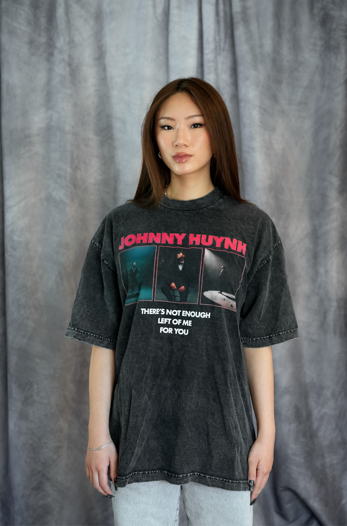 A young woman with straight brown hair stands before a gray curtain, wearing a faded black Johnny Huynh unisex relaxed fit mineral wash T-shirt with graphic prints and light jeans. She faces the camera neutrally.