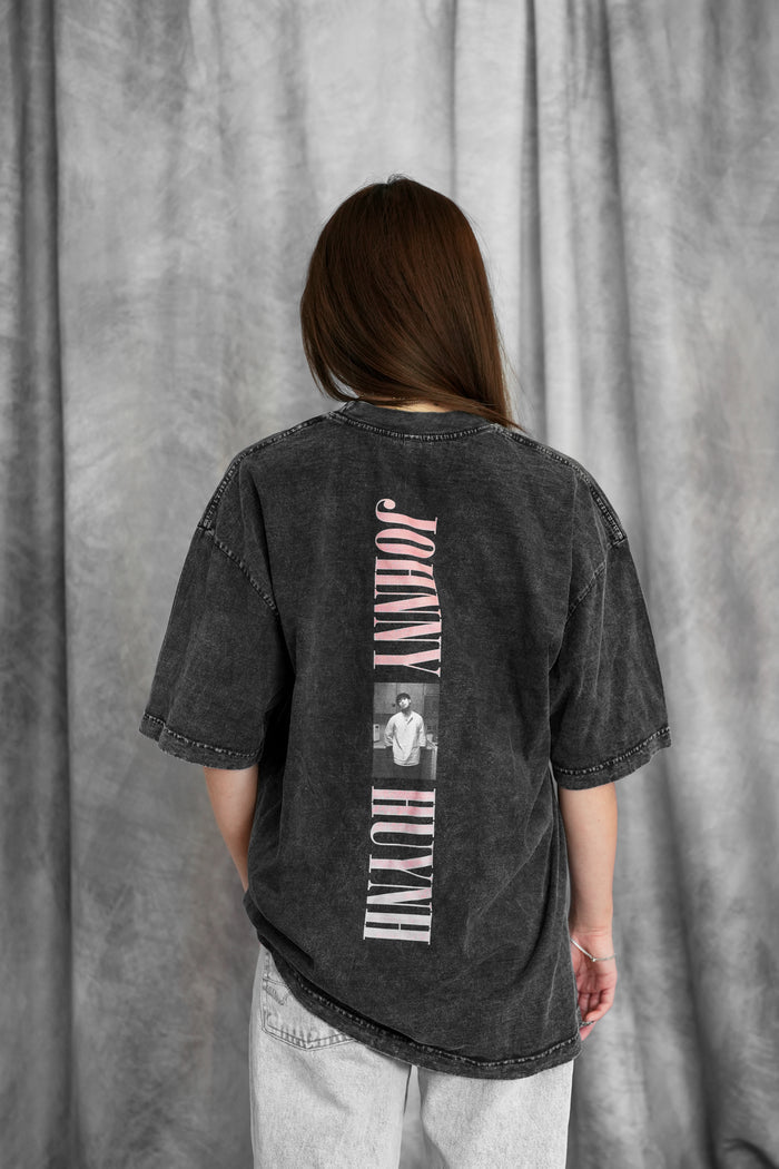 A person with long brown hair faces away, wearing a Johnny Huynh black mineral wash T-shirt with vertical text and a small grayscale photo on the back, paired with light gray jeans, set against draped gray fabric.