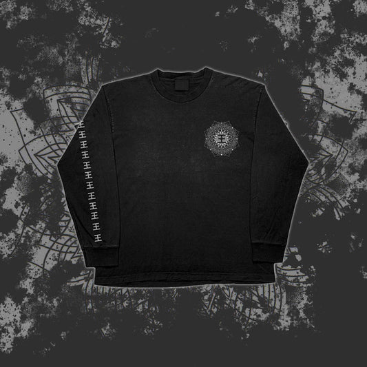 A black Reflections long sleeve shirt featuring a printed geometric mandala on the left chest and repeated white geometric symbols down the right sleeve, shown against a dark, grungy patterned background.