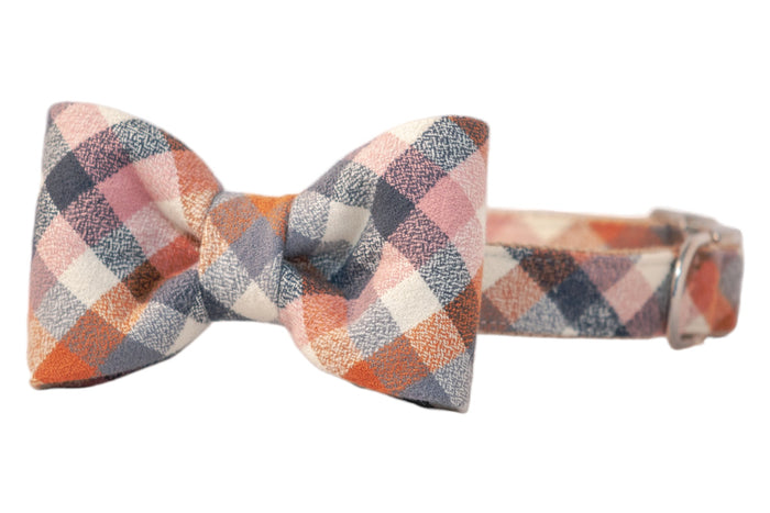 Close-up of a dog collar with an attached bow tie in a cozy autumn plaid flannel fabric. The pattern features small diagonal squares in warm tones. The collar includes a metal D-ring and a side-release buckle.