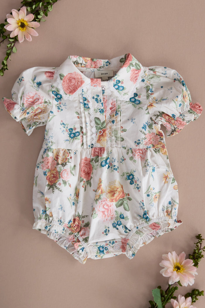 The Baby Sophia Romper in Countryside Peony Blossom by Ivy City Co features pastel floral cotton, short puffed sleeves, a gathered waist, pintuck chest detail, and snap crotch closures—shown on a beige background with flowers.
