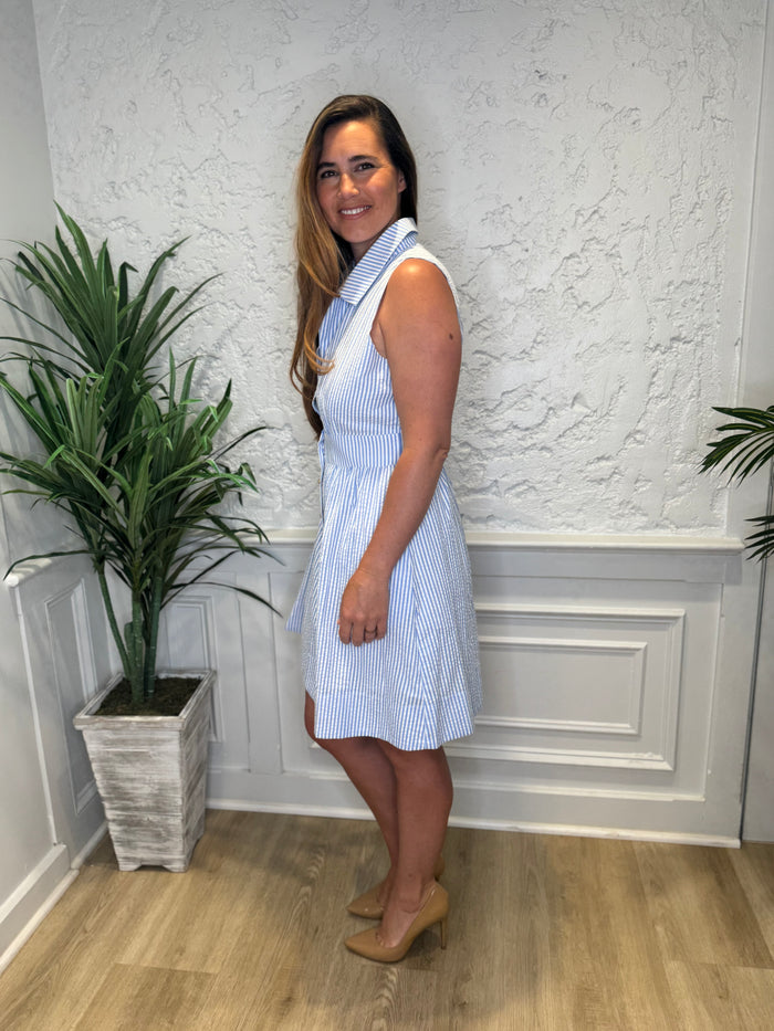 Side view of model in front of a white wall in the Duffield Lane Ridley Dress - Sky Seersucker. A knee length, seersucker dress with a collar, tailored waist, and gold buttons down the front