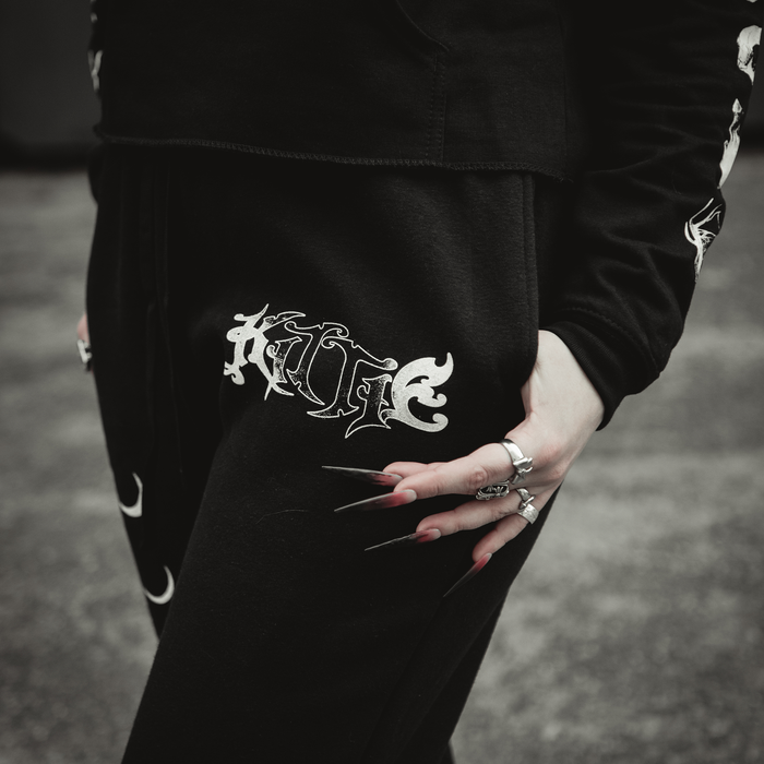 A person wearing black Kittie sweatpants with "KILLTEC" printed on them rests a ring-adorned hand with long pointed nails on their hip. Made from recycled polyester; background is blurred.