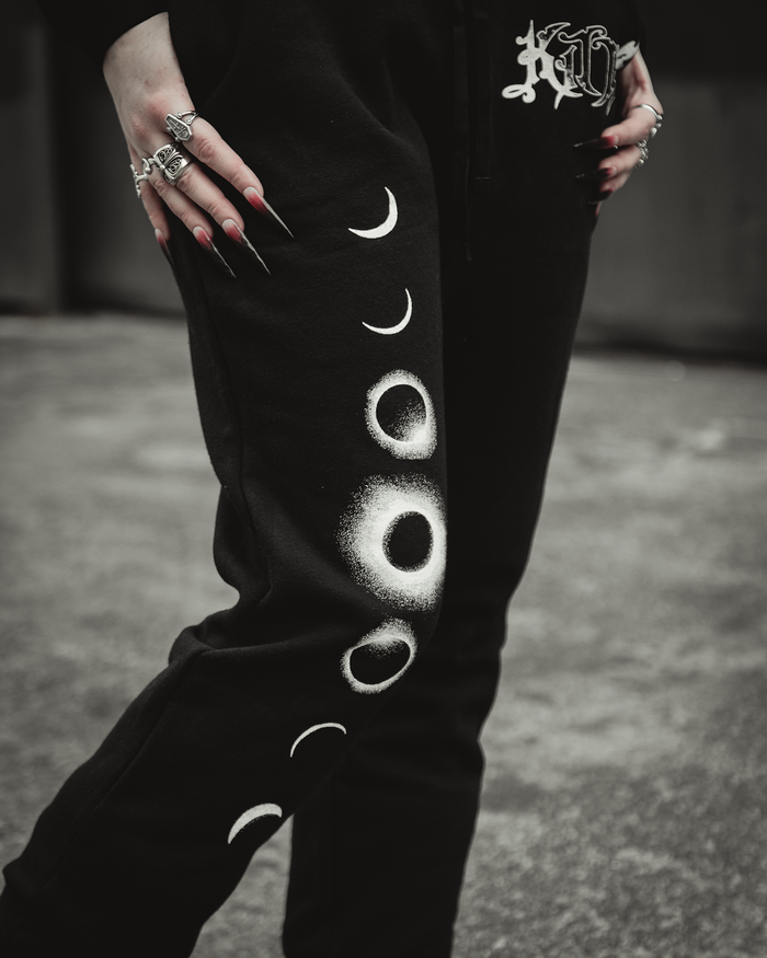 A person wears black Kittie sweatpants with white moon phases down one leg, multiple rings, long black nails, and a shirt or hoodie featuring a gothic Kittie logo. A blurred concrete background is visible.