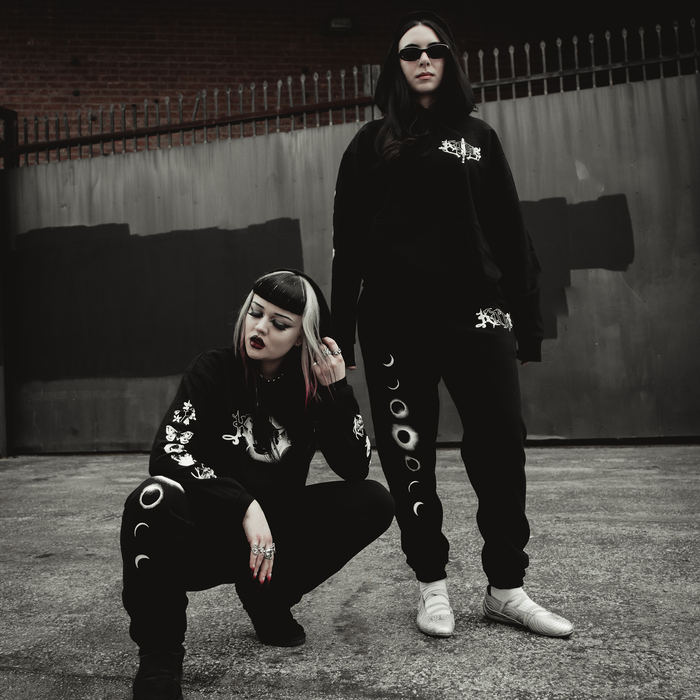 Two women in black Kittie relaxed-fit sweatpants with moon phase designs pose boldly in front of a metal gate, both sporting dark sunglasses. The gritty urban backdrop adds to the edgy vibe.