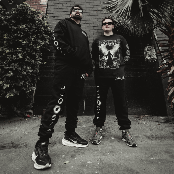 Two people stand confidently outdoors against a dark wall, both dressed in Kittie’s black moon-phase streetwear made from recycled polyester with bold graphic prints. Dark sunglasses and chunky sneakers complete their look, framed by palm leaves and brickwork.