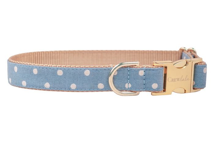 A light blue fabric dog collar with white polka dots, backed with beige nylon webbing and finished with a shiny gold buckle engraved with the brand name “Crewlala” and a matching gold D-ring.