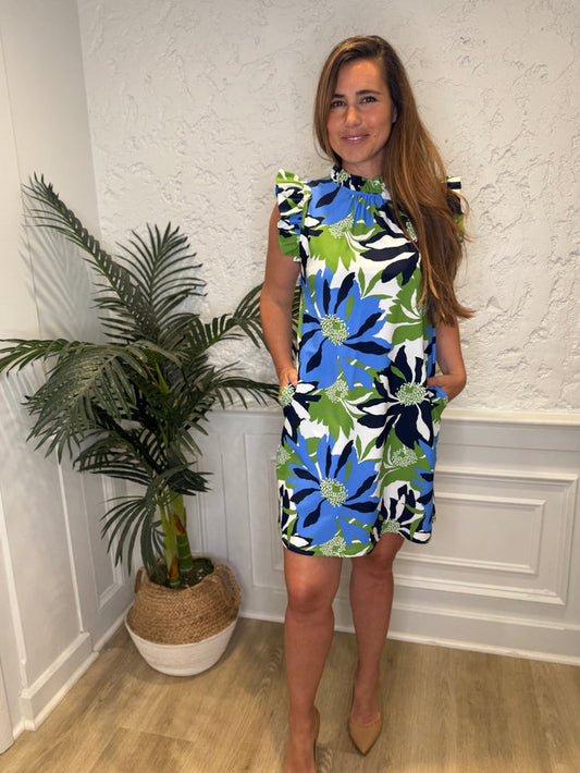Smiling model in front of a white wall with a plant in the corner in the Jude Connally Shari Dress - Wild Blossom Blue. A short dress with flutter sleeves and collar, with a blue, green, and navy floral pattern