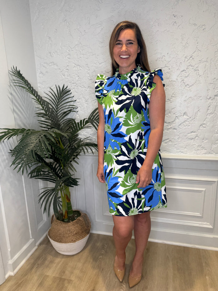 Smiling model in front of a white wall with a plant in the corner in the Jude Connally Shari Dress - Wild Blossom Blue. A short dress with flutter sleeves and collar, with a blue, green, and navy floral pattern