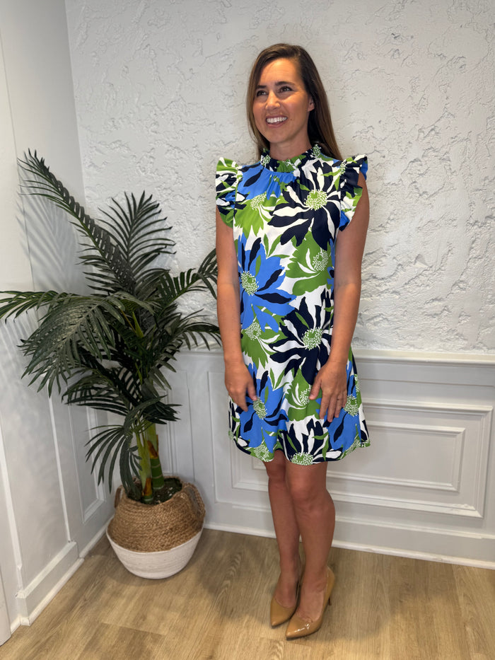 Smiling model in front of a white wall with a plant in the corner in the Jude Connally Shari Dress - Wild Blossom Blue. A short dress with flutter sleeves and collar, with a blue, green, and navy floral pattern