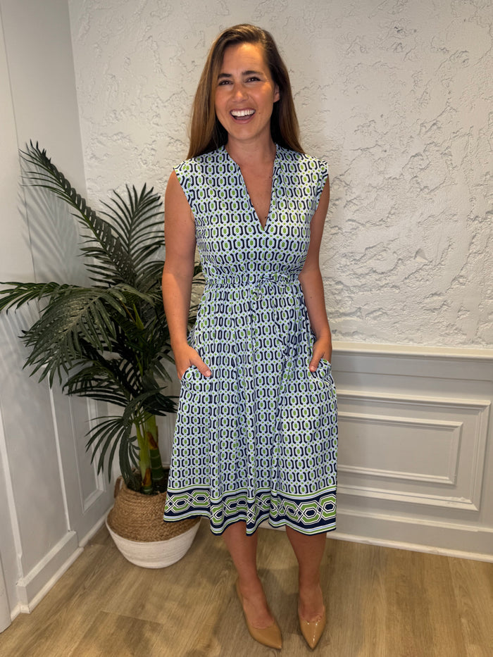 Smiling model in front of a white wall with a plant in the corner in the Jude Connally Tess Midi Dress - Terrace Geo Border Navy. A midi, v neck, sleeveless, drawstring waist dress with a white, green, and navy geometric pattern