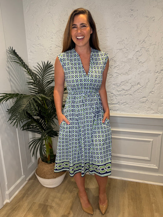 Smiling model in front of a white wall with a plant in the corner in the Jude Connally Tess Midi Dress - Terrace Geo Border Navy. A midi, v neck, sleeveless, drawstring waist dress with a white, green, and navy geometric pattern