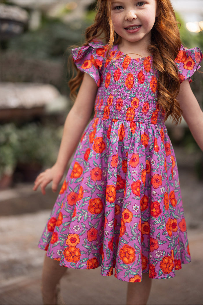 A young girl with long red hair smiles outdoors, wearing the Mini Hattie Dress in Thistle Flower Poppy Woodblock by Ivy City Co—a girls' cotton midi dress with puffy sleeves and a vibrant floral print on a purple background.