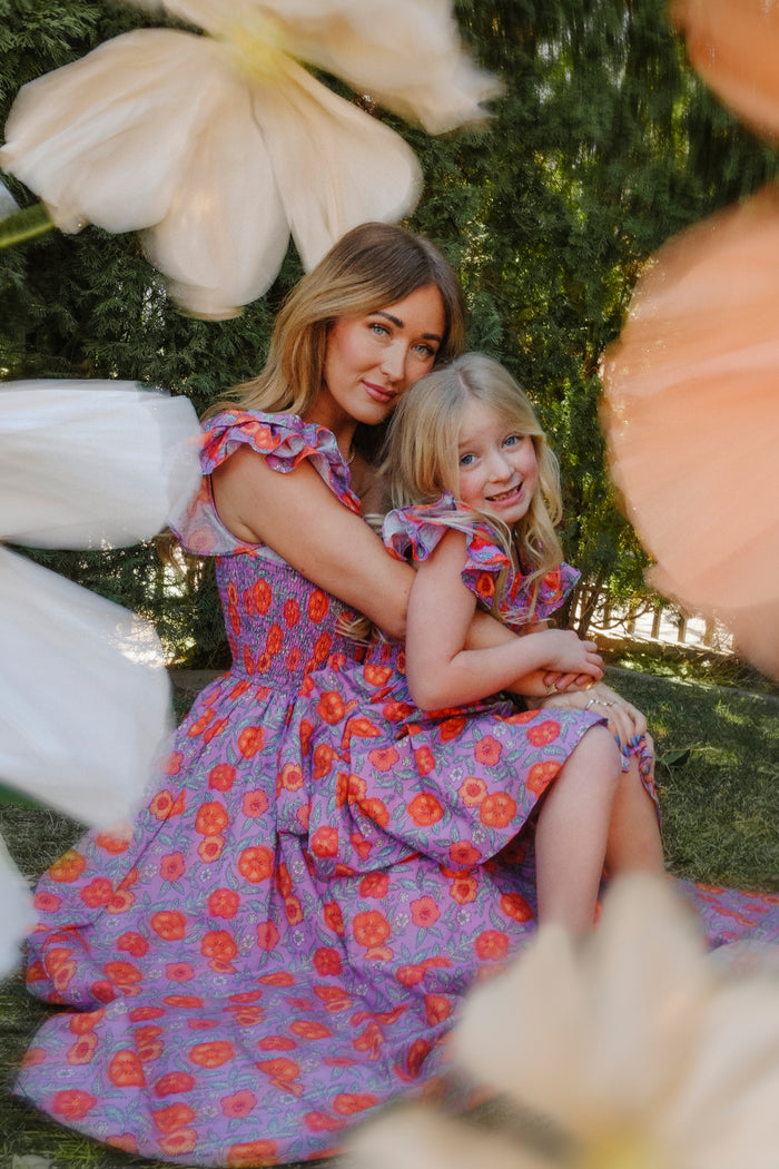 A woman and young girl smile as they sit outdoors, both wearing Ivy City Co's Mini Hattie Dress in Thistle Flower Poppy Woodblock. Surrounded by soft-focus flowers, they embrace in matching purple floral dresses with smocked bodices.