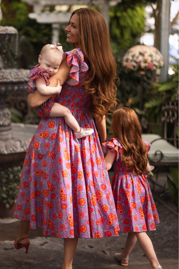 A woman with long brown hair wears the Ivy City Co Mini Hattie Dress in Thistle Flower Poppy Woodblock, holding a baby in a matching floral outfit while a young girl in similar attire walks beside them through a lush garden.