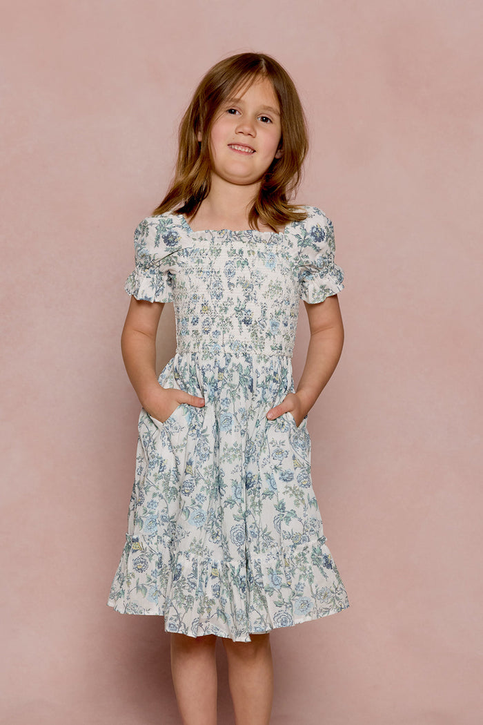 A young girl wearing the Ivy City Co Mini Valencia Dress in Ivory Climbing Vine Bloom, featuring short sleeves and pockets, stands against a pale pink background, smiling.