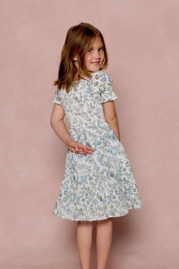 A young girl in the Mini Valencia Dress in Ivory Climbing Vine Bloom by Ivy City Co stands against a pink background, looking over her shoulder and smiling, with one hand behind her back and the other slightly raised.