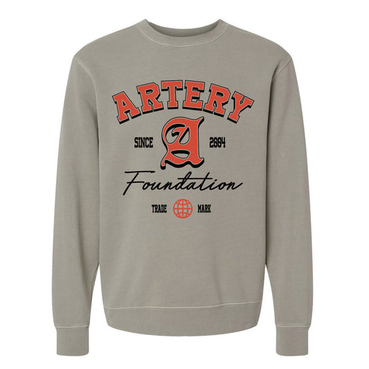 The Artery Foundation beige midweight pigment-dyed crewneck features bold red and black "ARTERY Foundation Since 2004" text, a large stylized A, and a small globe graphic above "TRADE MARK.