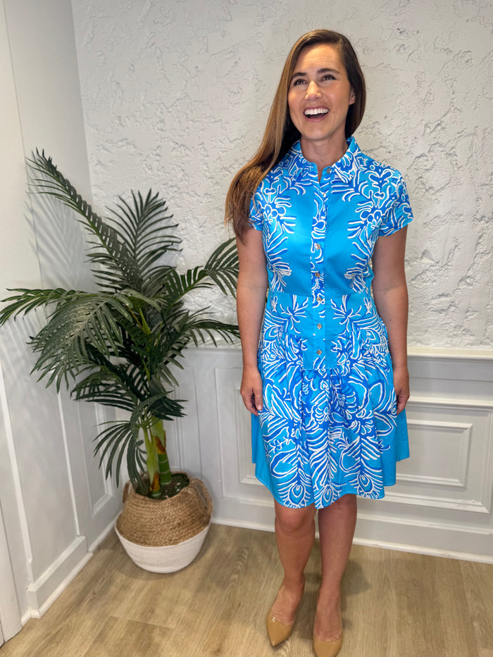 Smiling model in front of a white wall with a plant in the corner, in the Patty Kim Colette Dress - Aqua Rococo. A short sleeved mini dress with a drop waist and buttons down the front. A white and royal blue floral pattern on an aqua background