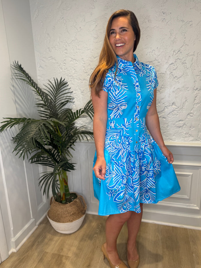 Smiling model in front of a white wall with a plant in the corner, in the Patty Kim Colette Dress - Aqua Rococo. A short sleeved mini dress with a drop waist and buttons down the front. A white and royal blue floral pattern on an aqua background