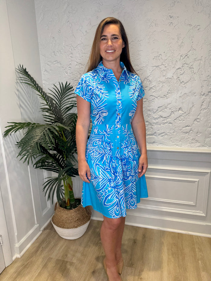 Smiling model in front of a white wall with a plant in the corner, in the Patty Kim Colette Dress - Aqua Rococo. A short sleeved mini dress with a drop waist and buttons down the front. A white and royal blue floral pattern on an aqua background