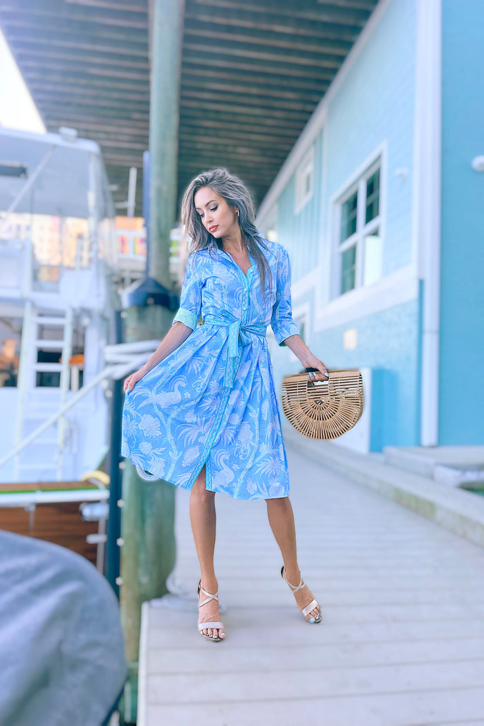 Model in front of a boat wearing a green blue and white patterned knee length dress. Cuffed sleeves, traditional collar, and belt
