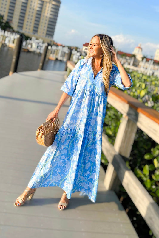Model on boardwalk in a tiered maxi dress with a light blue and white floral and bird pattern with touches of green. V-neck with traditional collar