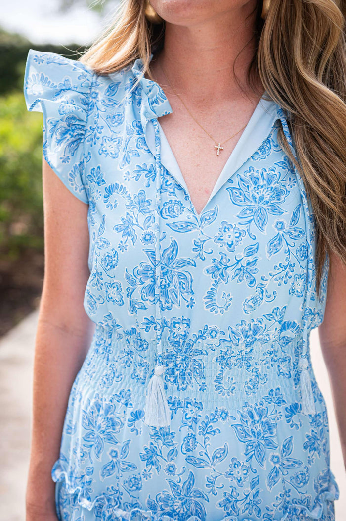 Model wearing a tiered maxi dress in a blue floral print with a ruched waist and flutter sleeves.Close up on details; ruched waist, flutter sleeves, and tassels