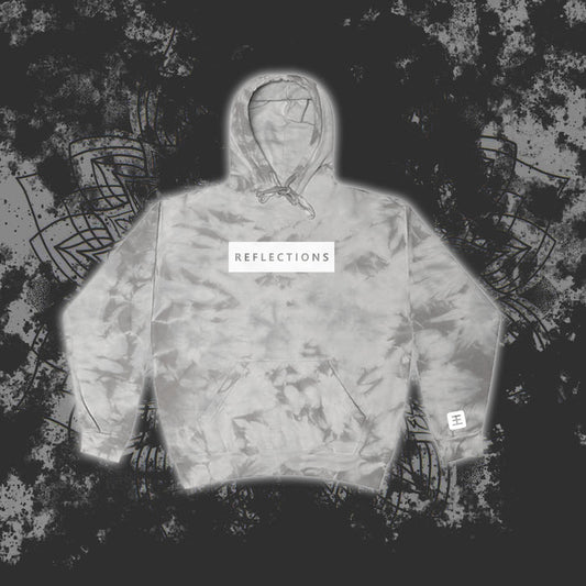 A gray Reflections tie-dye pullover hoodie with a white chest patch reading "REFLECTIONS" in black is showcased against a dark, abstract patterned background.