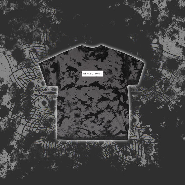 A charcoal black Reflections T-shirt with a white chest label and tie-dye pattern, displayed on a grunge-style gray and black abstract background with intricate line art.